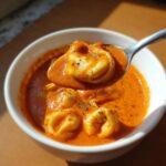 A spoonful of tortellini being lifted from a bowl of rich, creamy tomato tortellini soup, garnished with herbs.
