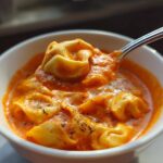 A spoonful of tortellini lifted from a bowl of rich, creamy tomato tortellini soup, garnished with herbs.
