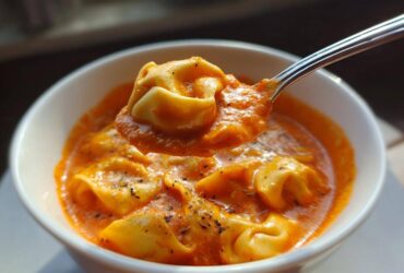 A spoonful of tortellini lifted from a bowl of rich, creamy tomato tortellini soup, garnished with herbs.