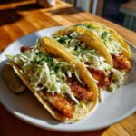 Three Crispy Baked Fish Tacos topped with bright green lime slaw and cilantro on a white plate.