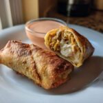 Two Crispy Baked Reuben Egg Rolls, one cut open showing melted cheese and filling, served with Thousand Island Dip.