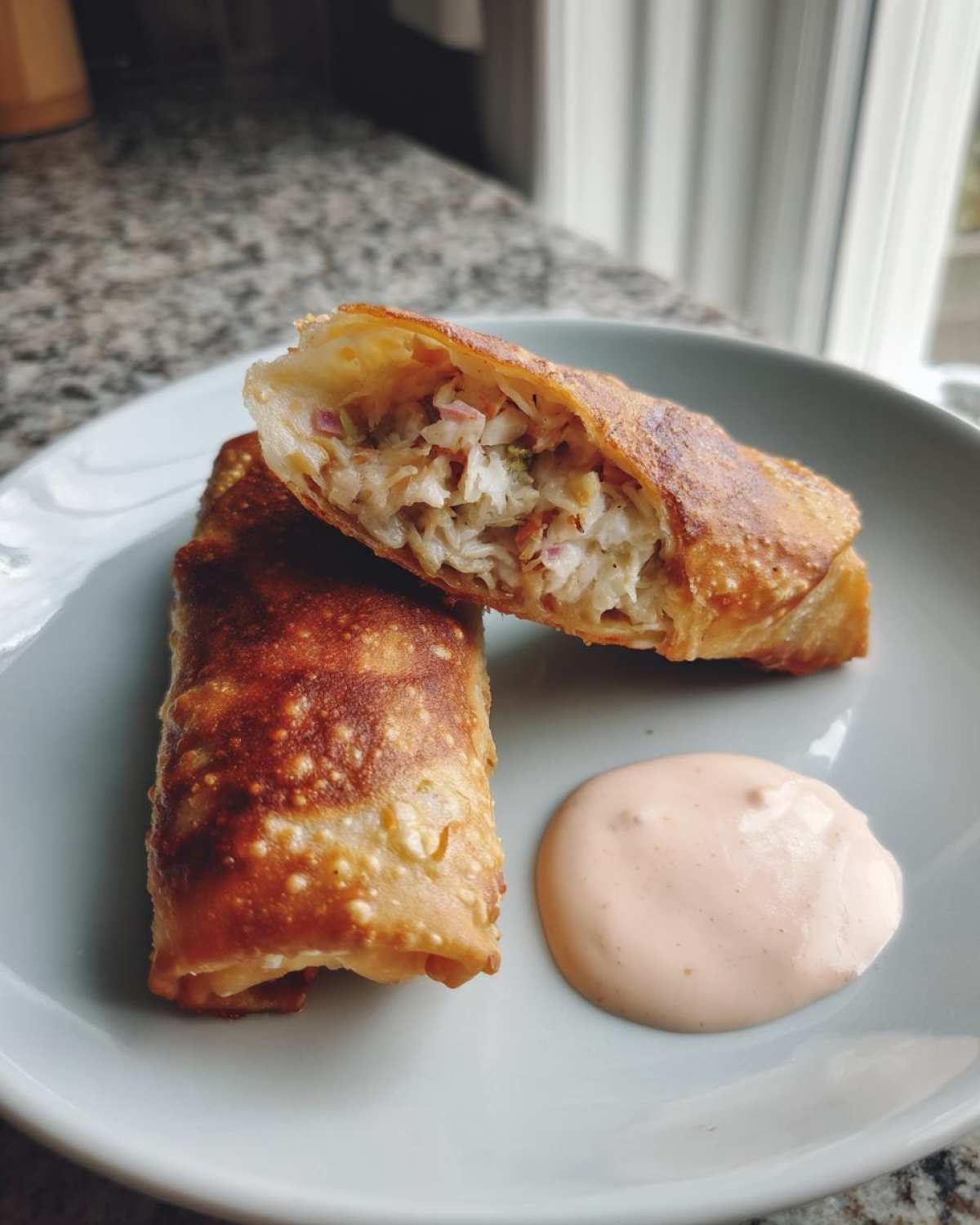 Two Crispy Baked Reuben Egg Rolls, one cut open showing the filling, served with Thousand Island Dip.