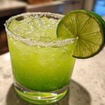 Close-up of a bright green Fresh Jalapeño Lime Margarita, served over ice with a salted rim and lime wheel garnish.