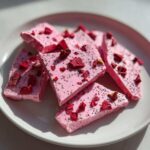 Several pieces of bright pink Frozen Strawberry Yogurt Bark topped with dried berry chunks and small seeds, arranged on a white plate.