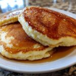 Close-up of three fluffy Gluten-Free Buttermilk Pancakes stacked and drizzled generously with golden syrup.