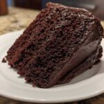 A close-up of a rich, dark slice of Gluten-Free Chocolate Cake covered in thick chocolate frosting on a white plate.