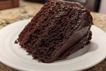 A close-up of a rich, dark slice of Gluten-Free Chocolate Cake covered in thick chocolate frosting on a white plate.