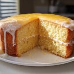 A two-layer Gluten-Free Lemon Coconut Cake with a slice removed, showing a thick lemon glaze and white frosting.
