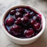Close-up of whole blueberries simmering in thick, dark purple liquid for Homemade Blueberry Syrup in a white bowl.