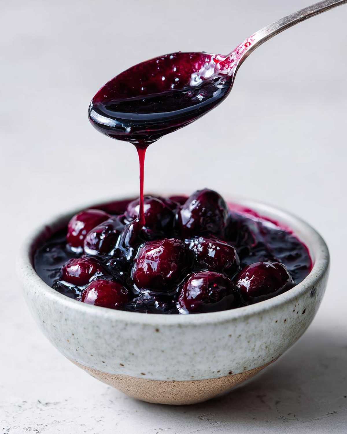 A spoonful of thick, dark Homemade Blueberry Syrup dripping over whole blueberries in a speckled ceramic bowl.