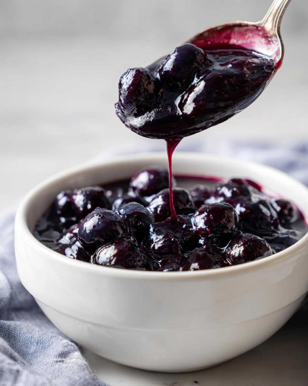 A spoonful of thick, rich Homemade Blueberry Syrup dripping back into a white bowl filled with cooked blueberries.