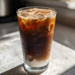 A tall glass of Iced Honey Almond Milk Latte showing layers of dark coffee, ice, and white almond milk at the bottom.