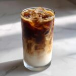 A tall glass of Iced Honey Almond Milk Latte showing rich dark espresso swirling into white almond milk over ice.