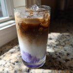 Close-up of a tall glass containing a layered Iced Lavender Latte with purple syrup, milk, espresso, and ice.