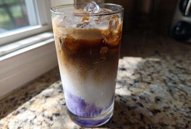 Close-up of a tall glass containing a layered Iced Lavender Latte with purple syrup, milk, espresso, and ice.