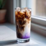 A tall glass of Iced Lavender Latte showing purple syrup, white milk, and dark coffee swirling over ice cubes.