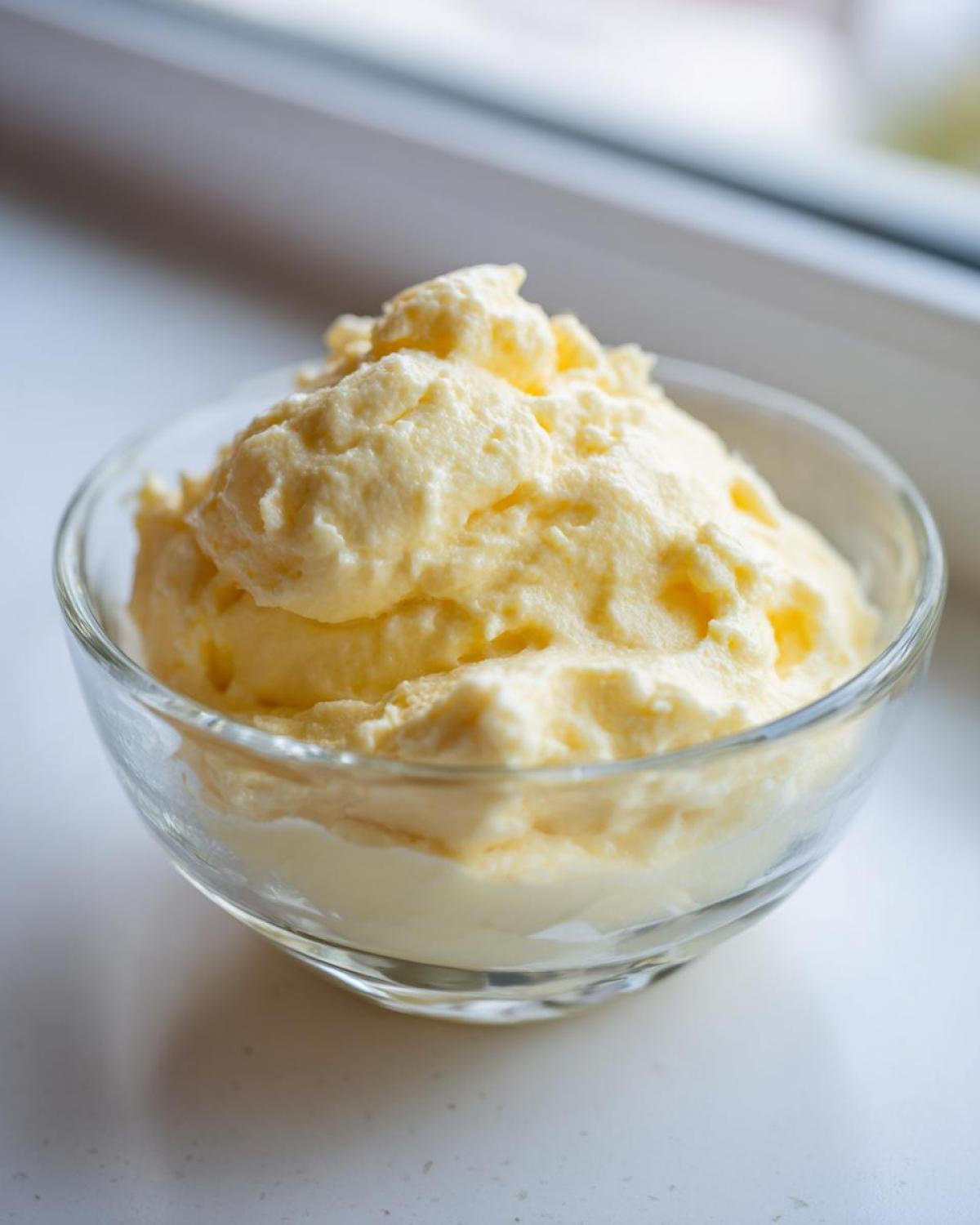 A serving of light yellow, fluffy Keto Lemon Cheesecake Mousse piled high in a small clear glass bowl.