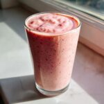 A tall glass filled with a thick, pink Keto Strawberry Cheesecake Smoothie, sitting on a bright windowsill.