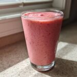 A tall glass filled with a thick, pink Keto Strawberry Cheesecake Smoothie sitting on a speckled countertop near a window.