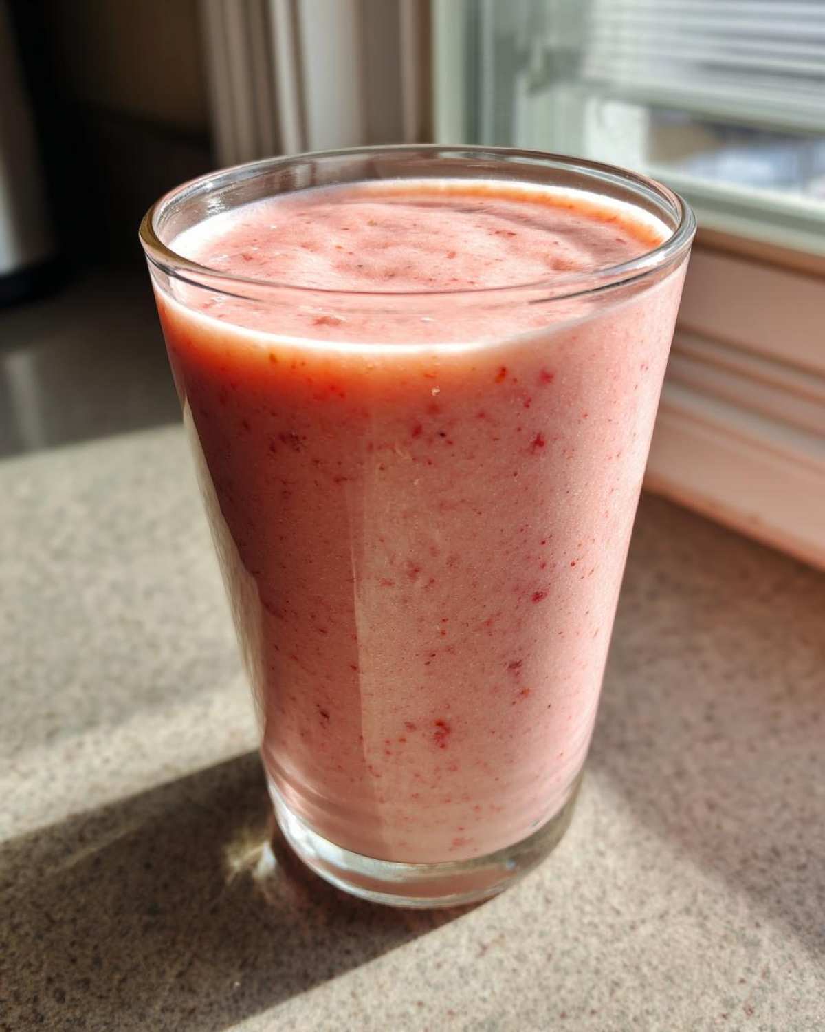 A tall glass filled with a thick, pink Keto Strawberry Cheesecake Smoothie, sitting on a countertop in natural sunlight.