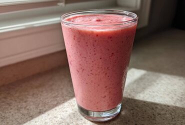 A tall glass filled with a thick, pink Keto Strawberry Cheesecake Smoothie sitting on a speckled countertop near a window.