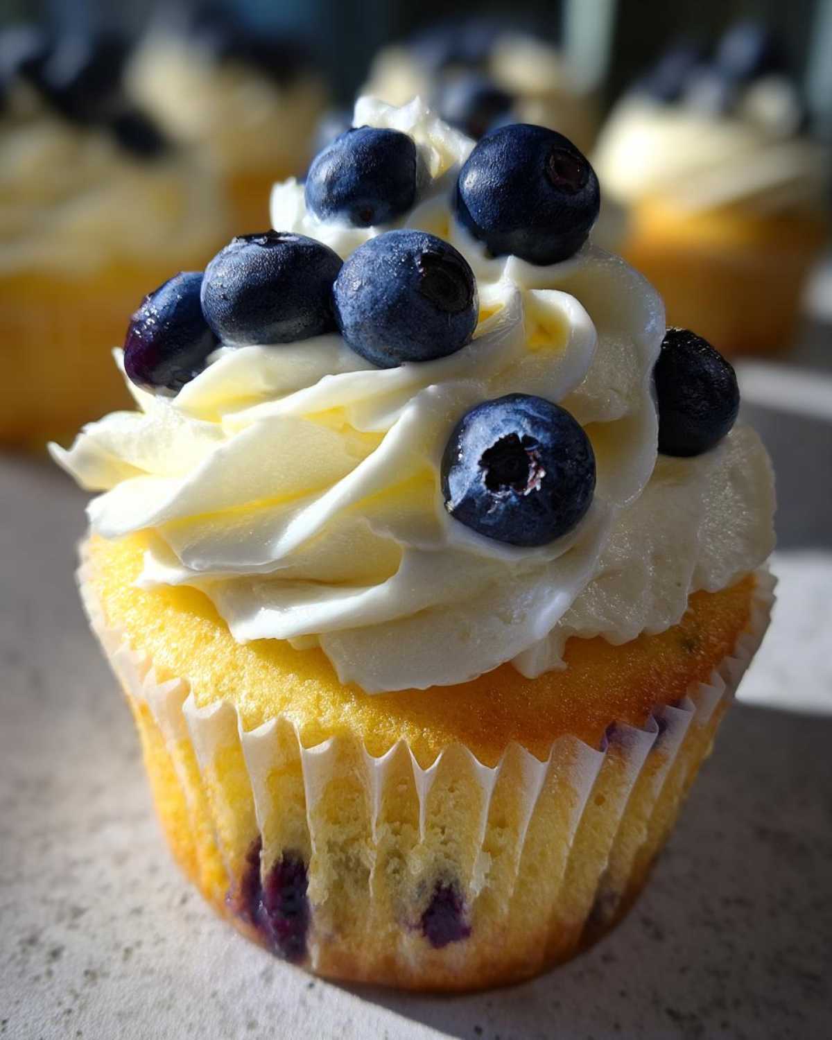 A close-up of a Lemon Blueberry Cupcake topped with swirls of cream cheese frosting and fresh blueberries.