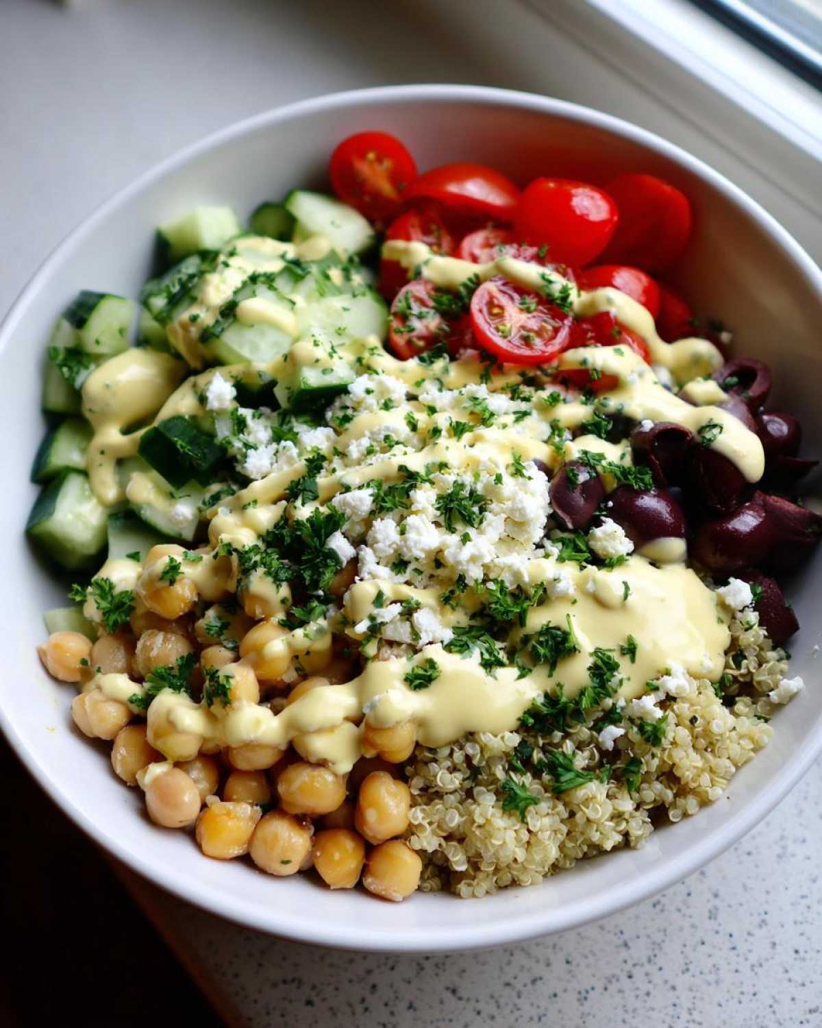 Close-up of a Mediterranean Quinoa Bowl topped with chickpeas, feta, tomatoes, cucumbers, and lemon tahini sauce.