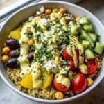 Close-up of a vibrant Mediterranean Quinoa Bowl topped with feta, tomatoes, cucumbers, and lemon tahini sauce.