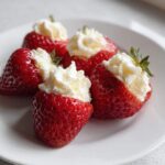 Four bright red strawberries filled with creamy white No-Bake Cheesecake Stuffed Strawberries filling on a white plate.