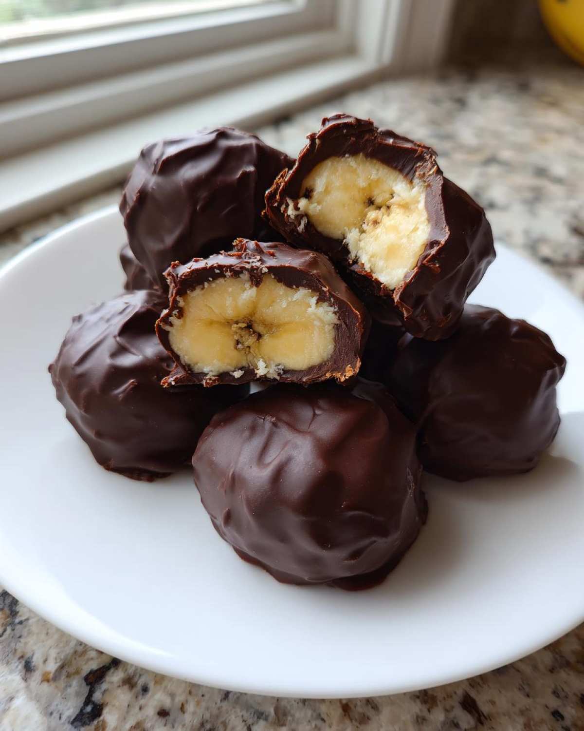 A pile of No-Bake Chocolate Covered Banana Bites on a white plate, one is cut open showing banana inside.