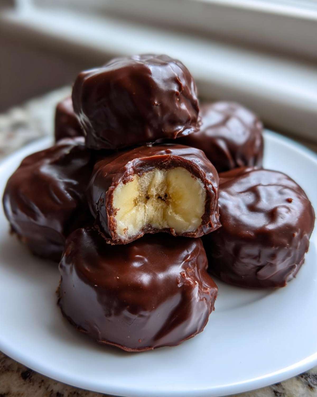 A stack of shiny, dark chocolate covered banana bites, with one bitten into revealing the frozen banana center.