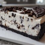 A thick slice of No-Bake Oreo Cheesecake featuring a dark cookie crust and creamy filling studded with chocolate pieces.