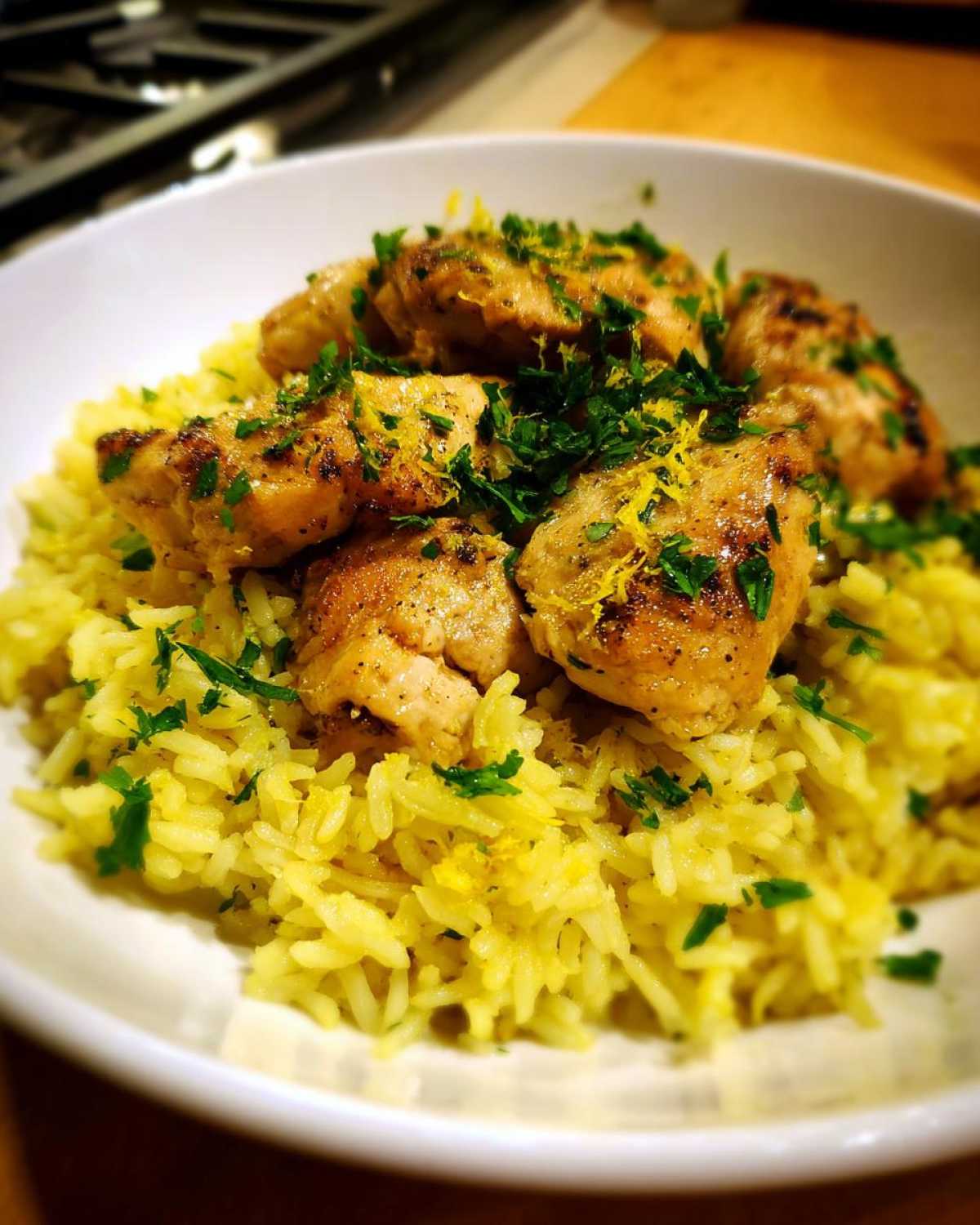 Close-up of a white bowl filled with yellow lemon rice topped with browned chicken pieces and fresh parsley.