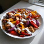 Close-up of Sheet Pan Chicken Shawarma with roasted peppers and onions, drizzled generously with garlic yogurt sauce.