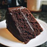 A moist, dark slice of Gluten-Free Chocolate Cake with rich chocolate frosting on a white plate.