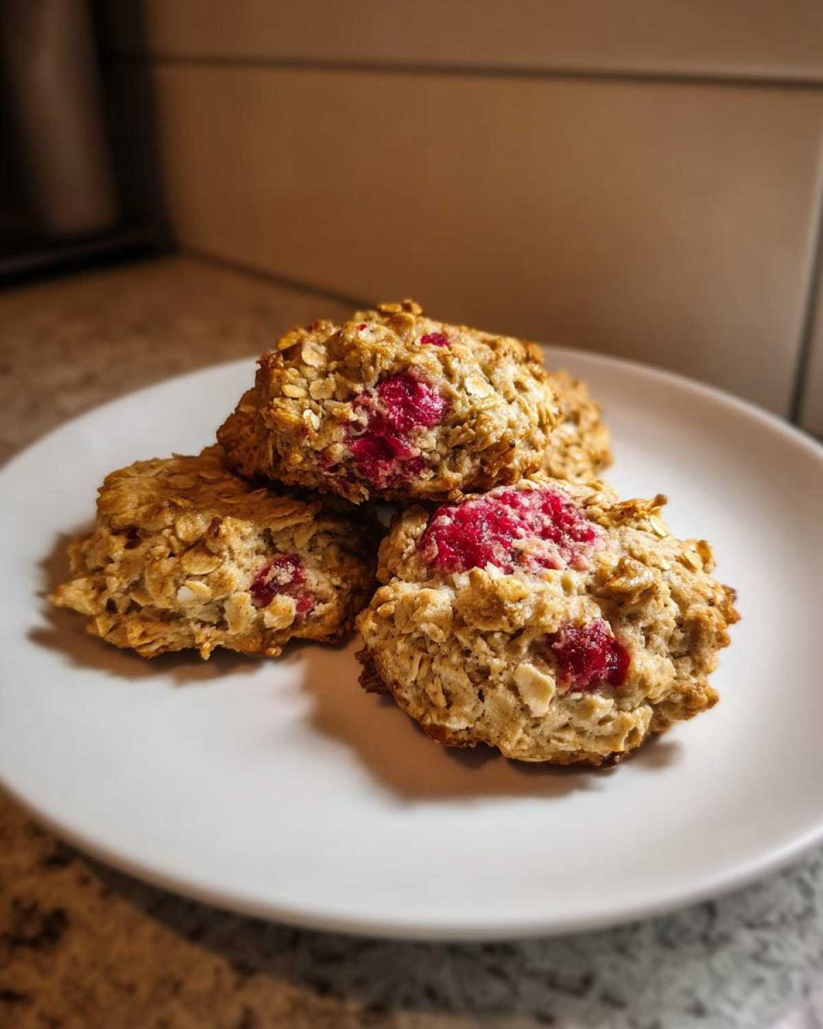 Three golden Strawberry Oatmeal Breakfast Cookies piled on a white plate, showing visible oats and bright red strawberry pieces.