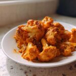 A white plate filled with bright orange florets of Vegan Cauliflower Tikka Masala, sitting on a speckled countertop.