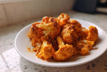 A white plate filled with bright orange florets of Vegan Cauliflower Tikka Masala, sitting on a speckled countertop.
