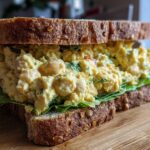 Close-up of a thick Vegan Chickpea Salad Sandwich overflowing with creamy filling between two slices of seeded whole grain bread.