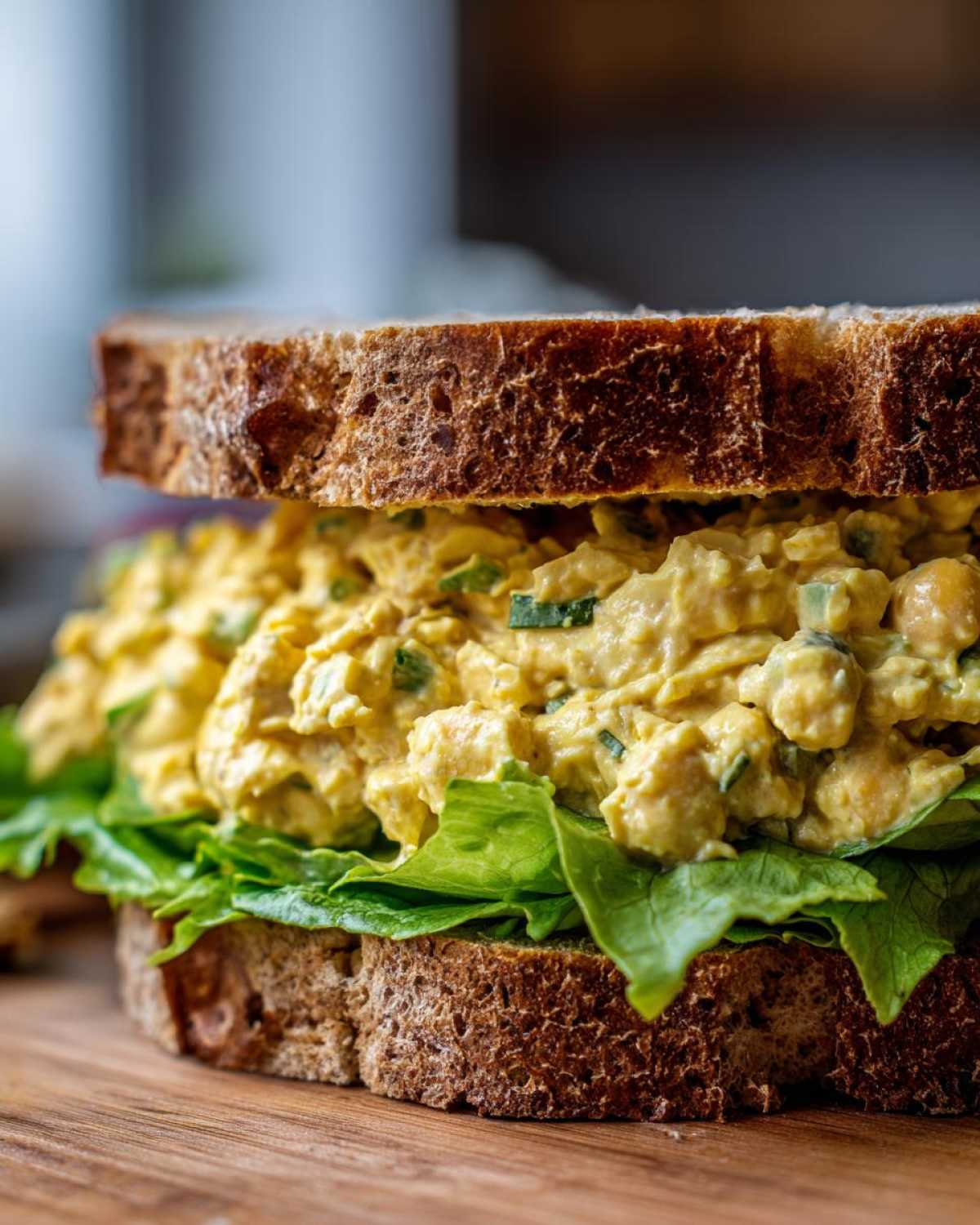 Close-up of a delicious Vegan Chickpea Salad Sandwich piled high with yellow chickpea salad and crisp lettuce on dark wheat bread.
