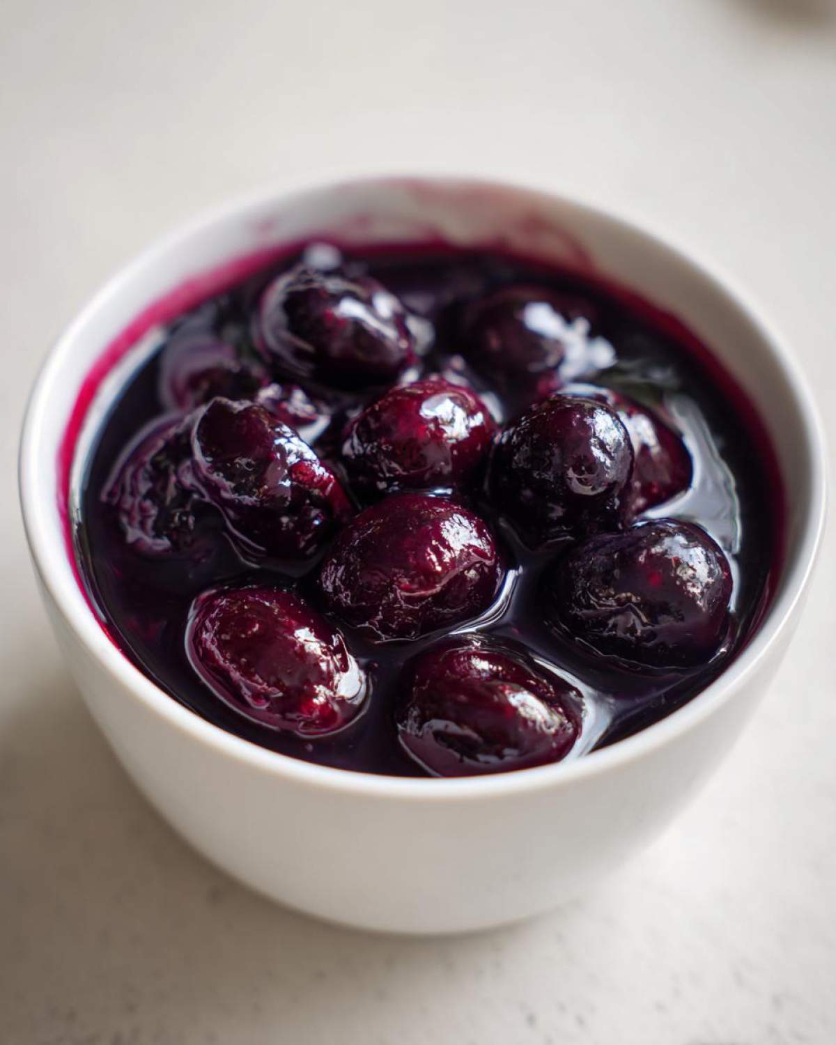 Close-up of plump, dark blueberries swimming in rich liquid for homemade blueberry syrup in a white bowl.