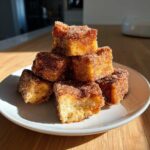 A stack of golden Air Fryer Crispy Cinnamon French Toast Bites coated heavily in cinnamon sugar on a white plate.