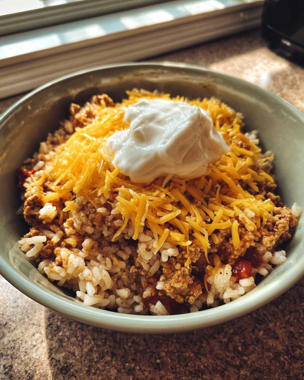 A close-up of a bowl filled with rice, seasoned ground turkey, shredded cheddar cheese, and a dollop of sour cream, representing Air Fryer Crispy Turkey Taco Bowls.