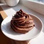 A rich swirl of homemade Chocolate Buttercream Frosting piped onto a small white plate, highlighted by natural sunlight.