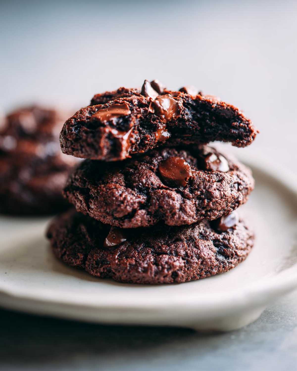A stack of three rich, dark Chocolate Chocolate Chip Cookies, with the top one broken open showing melted chocolate chips.