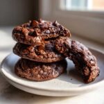 A stack of three fudgy Chocolate Chocolate Chip Cookies on a speckled plate, with one cookie broken open.