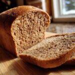 A loaf of freshly baked Copycat Dark Brown Honey Wheat Bread, partially sliced on a wooden board, highlighted by sunlight.