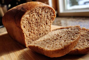 A loaf of freshly baked Copycat Dark Brown Honey Wheat Bread, partially sliced on a wooden board, highlighted by sunlight.