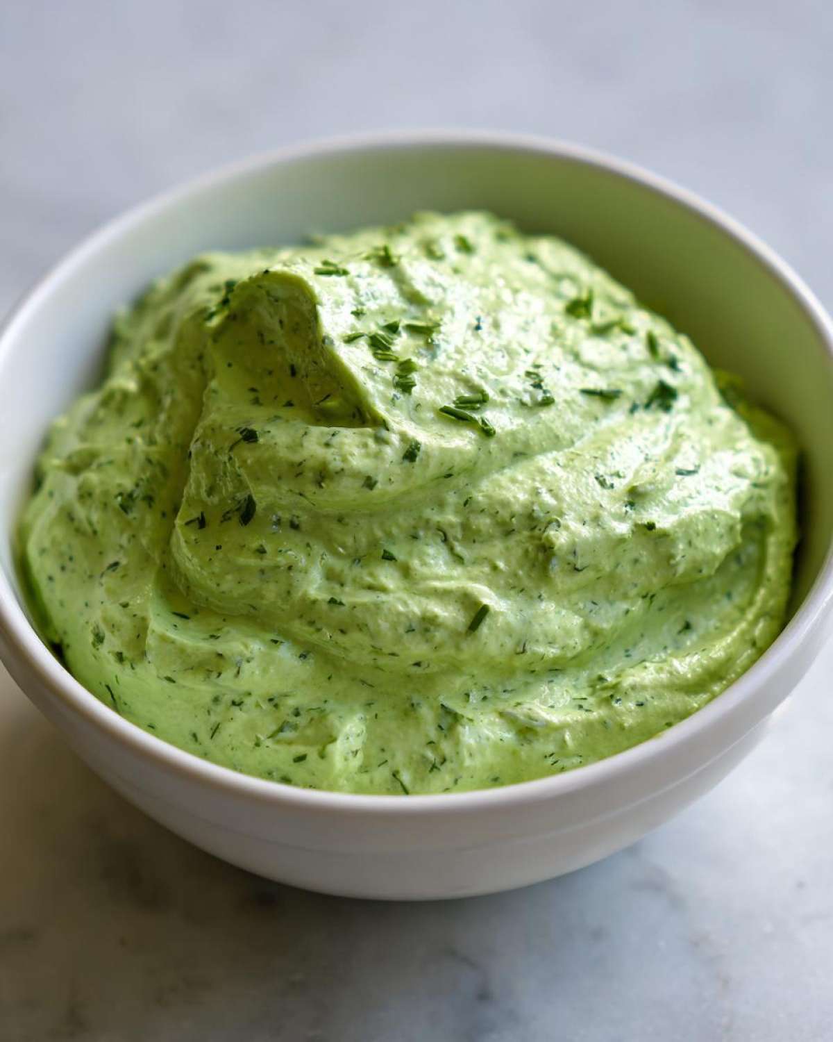 Close-up of thick, vibrant green Copycat Green Goddess Ranch Dressing topped with fresh herbs in a white bowl.