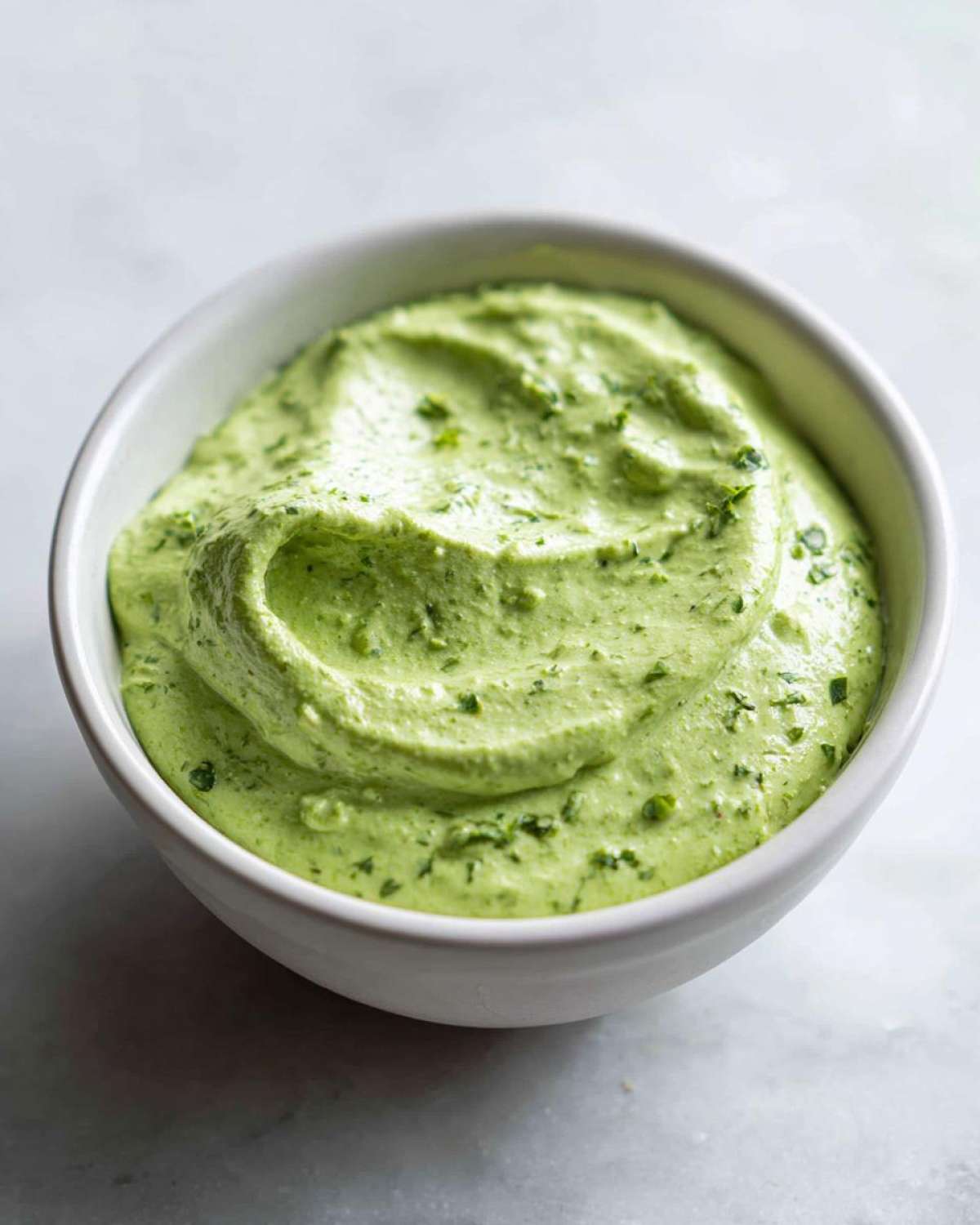 Close-up of a white bowl filled with thick, vibrant green Copycat Green Goddess Ranch Dressing flecked with herbs.